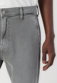 Γκρι τζιν με ίσια γραμμή, θηλιές ζώνης, μεταλλικό κουμπί και διακριτές ραφές. Το ύφασμα έχει λεία υφή.