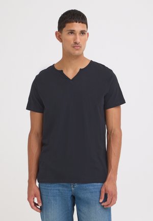 Homme portant un t-shirt noir uni à manches courtes avec un col en V et un jean bleu, debout devant un fond blanc.