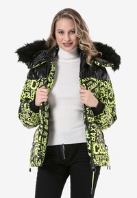 Gevoerde zwart en neon groene jas met grafische tekst, bontvoering in de capuchon, ritszakken en ribgebreide manchetten, gedragen over een coltrui.