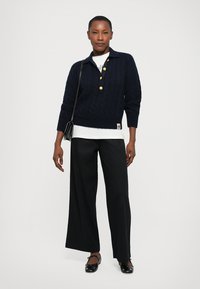 Femme portant un pull en maille torsadée bleu marine avec des boutons dorés, un sous-vêtement blanc, un pantalon large noir, un sac à main noir et des ballerines noires.