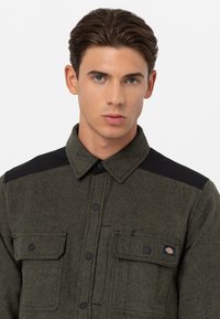 Giacca camicia in lana verde oliva con pannelli neri sulle spalle, bottoni a pressione e due tasche anteriori con un logo del marchio sulla tasca sinistra.