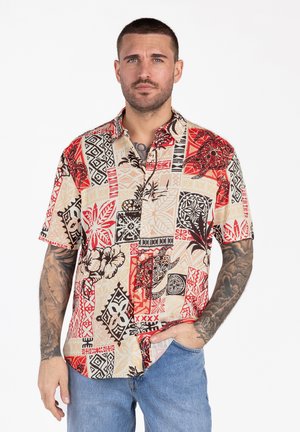 Man met kort haar die een rood, beige en zwart overhemd met korte mouwen en blauwe spijkerbroek draagt, staand met één hand in de zak.