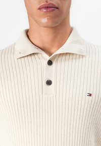Gros plan sur une personne portant un t-shirt côtelé crème avec deux boutons noirs et un petit logo de drapeau rouge, blanc et bleu brodé sur la poitrine.