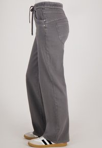 Grijze wijde broek met elastische tailleband en trekkoord, gedragen met witte sneakers met zwarte strepen en gumrubberen zolen.