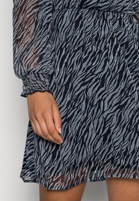 Robe bleu marine en tissu transparent texturé avec un motif zébré en zigzag. Poignets élastiqués et coupe décontractée à la taille.