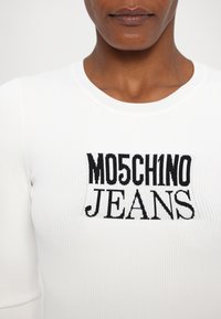 Bela rebrasta pletenina z črnim grafičnim napisom "MO5CH1NO JEANS," ki izpostavlja logotip blagovne znamke.
