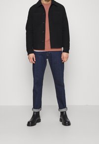 Chaqueta negra de botones sobre una sudadera color óxido, combinada con jeans de mezclilla azul y botines negros. Texturas de tela suaves, ajuste casual.