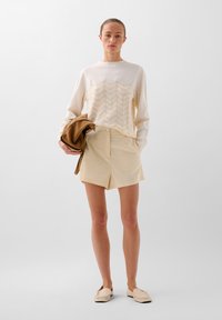 Cremefarvet sweater med bølget tekstur, parret med lysebeige shorts. Modellen holder en brun accessory. Let, blød stof; afslappet pasform.