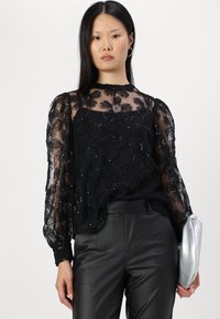 Mujer vistiendo una blusa de encaje negro floral transparente con lentejuelas, pantalones de cuero negro y sosteniendo un bolso de mano plateado frente a un fondo liso.