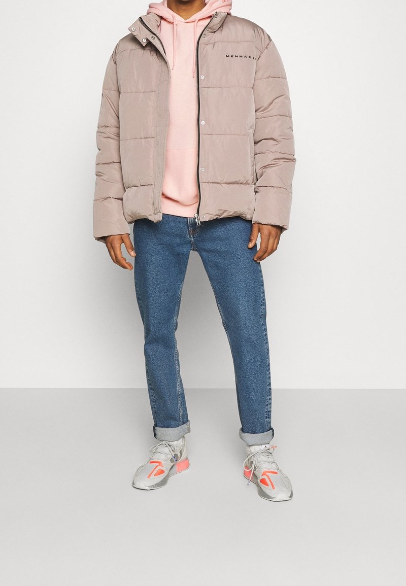 Chaqueta acolchada color beige claro con cierre de cremallera, con detalle de logotipo y capucha fija; combinada con una sudadera rosa claro y jeans azules.