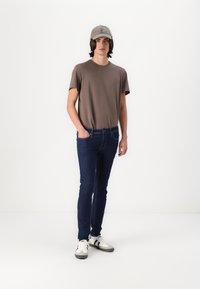 Szürke, rövid ujjú póló, sötétkék, slim-fit farmernadrág, szürke sapka logóval, és fehér sportcipő fekete részletekkel. A modell áll, keze a zsebében.