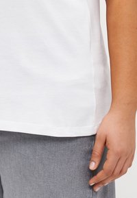 White crew-neck t-shirt displayed with short sleeves, side slit details, and a smooth texture, paired with gray textured pants.