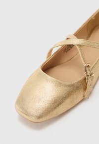 Kultaiset ballerinat, joissa on teksturoitu metallipinta, neliön muotoinen kärki, ristihihnadesign ja pieni hopeinen solkipisto. Sujuva sisävuori.