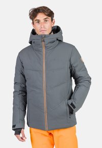 Graue gepolsterte Jacke mit hohem Kragen, Reißverschluss mit orangefarbenem Akzent und taillierten Bündchen; kombiniert mit leuchtend orangefarbenen Hosen.