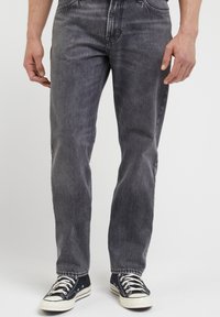 Person som bär urtvättade svarta raka jeans och svarta låga sneakers med vita snören, stående mot en plain bakgrund.