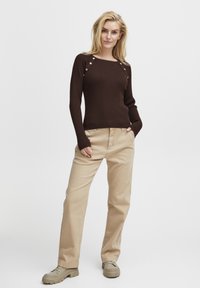 Maglione marrone a coste con dettagli di bottoni sulle spalle, abbinato a pantaloni beige a gamba dritta e scarpe chunky beige. Look semplice e dalla vestibilità rilassata.