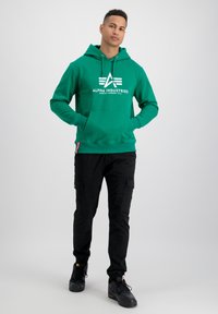 Sudadera verde con logo blanco, bolsillo tipo canguro, capucha con cordón, combinada con pantalones cargo negros y zapatillas negras. Fondo liso.