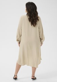 Beige lange mouw jurk met een zachte textuur, losse pasvorm en een gebogen zoomlijn. Beschikt over knoopmanchetten en valt tot halverwege het scheenbeen.