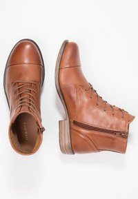 Botas de cuero marrón hasta el tobillo con punta redondeada, cordones delanteros y cremallera lateral. Acabado texturizado y detalle de tacón de madera apilada.
