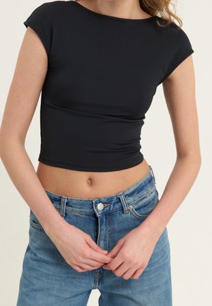 Vrouw draagt een nauwsluitend zwart cropped top met korte mouwen en een high-waist blauwe jeans, handen voor zich samengevouwen.