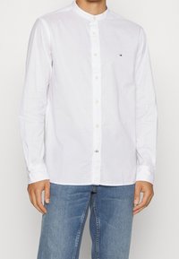 Camicia bianca a maniche lunghe con colletto alla coreana, chiusura con bottoni frontale e piccolo dettaglio del logo sul lato sinistro del petto. Tessuto di cotone liscio.