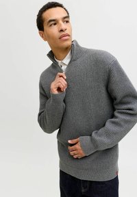 Vīrietis pelēkā, ribotā, ar rāvējslēdzēju apģērbtā džempera un tumšās biksēs, tur rāvējslēdzēju netālu no zoda, nedaudz raugoties uz sāniem.