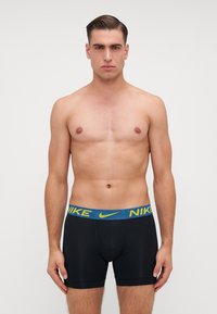 Fekete elasztikus anyagú boxer sort kék és sárga mintás derékrésszel, amelyen a "NIKE" logó látható. Sima textúra, testhezálló kivitel.