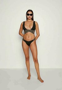 Olivgrön bikini med ett geometriskt guldmönster över bröstet och midjebandet. Slank design med en djup uringning och minimal täckning.