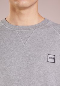 Sudadera gris con cuello y costuras de los hombros acanaladas, con una etiqueta rectangular negra y gris que muestra "BOSS" y "HUGO BOSS."