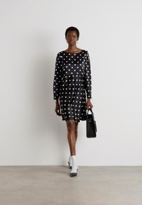 Abito nero con pois, maniche lunghe, tessuto texturizzato, gonna a campana, abbinato a una piccola borsa nera e stivaletti bianchi.