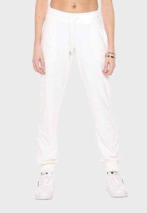Femme portant un pantalon de jogging blanc avec taille à cordon, des baskets blanches et des bracelets en or au poignet droit, debout sur un fond clair.
