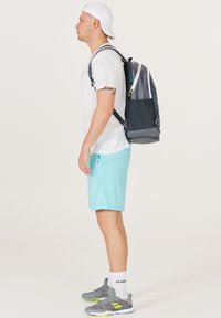 Mochila en malla gris y negra, con un logo triangular, correas ajustables y un asa en la parte superior. El modelo lleva una camisa blanca y pantalones cortos azul claro.