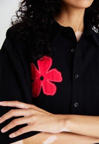 Sort button-up skjorte med fremtrædende rød blomsterbroderi på venstre skulder. Materialet ser tekstureret ud med en afslappet pasform.