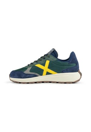 Sporty sneaker i mørkeblå, grøn og grå med et skarpt gult X på siden og en hvid sål med brun mønster.