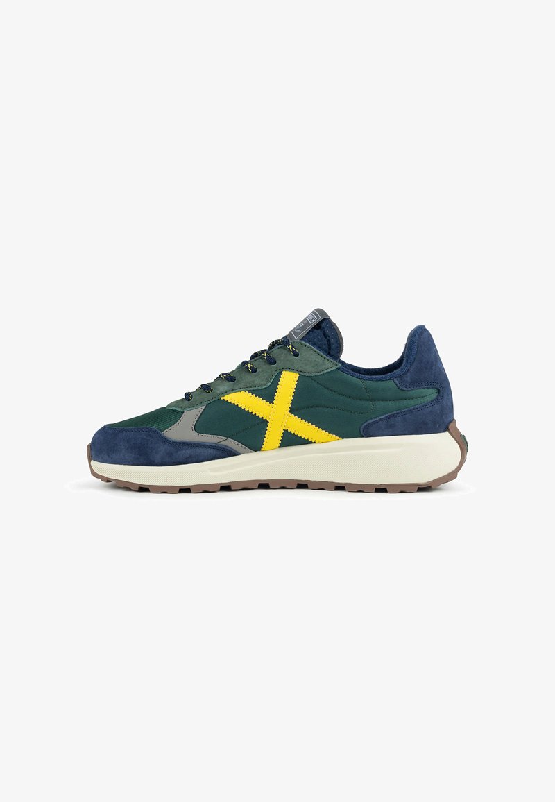 Baskets sportives bleu marine, vert et gris avec un X jaune vif sur le côté et une semelle blanche avec un profil marron.