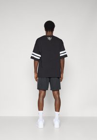 Svart oversized t-skjorte med hvite striper på ermene, logo på baksiden, sammen med mørke shorts og hvite joggesko.