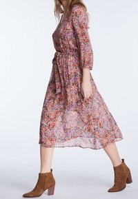 Maxikleid mit Blumenmuster in Pink, Orange und Lila; hat einen V-Ausschnitt, eine taillierte Taille und Dreiviertelärmel. Kombiniert mit braunen Stiefeletten.