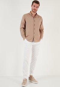 Camicia button-up color marrone chiaro con colletto, abbinata a pantaloni bianchi e sneaker beige. La camicia presenta maniche lunghe e una texture sottile.