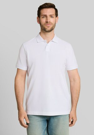 Mann mit kurzen braunen Haaren und Bart, der ein weißes kurzärmeliges Poloshirt und hellblaue Jeans vor einem schlichten Hintergrund trägt.