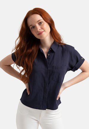 Donna con lunghi capelli rossi che indossa una camicia blu navy a maniche corte con bottoni e pantaloni bianchi, mani sui fianchi, leggermente sorridente, su uno sfondo uniforme.