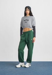 Γκρι κοντή φούτερ με μαύρο εκτύπωση «Nike 1972», συνδυασμένη με πράσινα παντελόνια jogging και λευκά αθλητικά παπούτσια με πράσινες λεπτομέρειες.