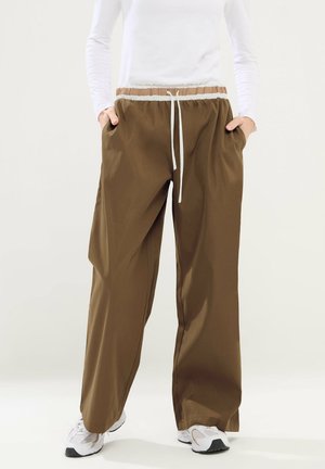 Osoba nosząca luźne brązowe spodnie z białym sznurkiem i kieszeniami, w połączeniu z białymi trampkami i białą bluzką z długim rękawem.