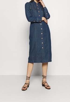 Kvinde iført en langærmet knap-op denim midi-kjole kombineret med sorte sandaler med blokhæl og stropper, poserer med én hånd nær ansigtet.