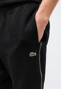 Άτομο που φοράει μαύρο παντελόνι με το χέρι στην τσέπη, με μικρό πράσινο λογότυπο κροκοδείλου και πλαϊνή ρίγα με κείμενο σε λευκό φόντο.