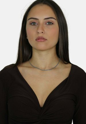 Giovane donna con capelli lunghi e scuri che indossa una maglietta nera a scollo a V e una sottile collana d'argento, rivolta verso avanti con un'espressione neutra.