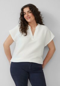 Witte, gestructureerde top met een V-hals en korte mouwen, met een lossere pasvorm. Gecombineerd met donkerblauwe broeken.