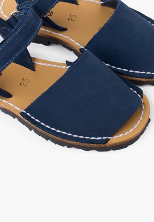 Sandales bleu marine avec une tige en cuir lisse, bout carré et coutures blanches sur une semelle intérieure beige. Dotées d'une semelle extérieure en caoutchouc noir robuste.