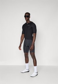 Svart kortermet sykkeldrakt med glidelås foran, kombinert med tettsittende mørke shorts. Brukt med hvite sokker og hvite sykkelsko.