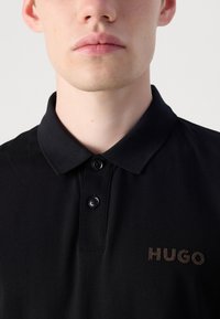 Juodas polo marškinėliai su tekstūruotu audiniu, dviem mygtukais ant apykaklės ir rudai siuvinėtu "HUGO" logotipu kairėje pusėje.