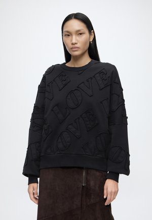 Femme aux cheveux longs et raides portant un pull noir oversize avec des lettres "LOVE" texturées et une jupe marron foncé avec une fermeture éclair.